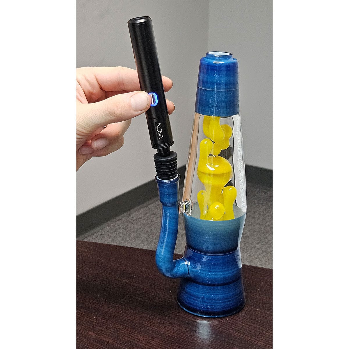 Handheld top-down view of a black Cipher NOVA Water Pipe Adapter being attached to the blue glass water pipe, shown against a gray wall and dark wooden table.