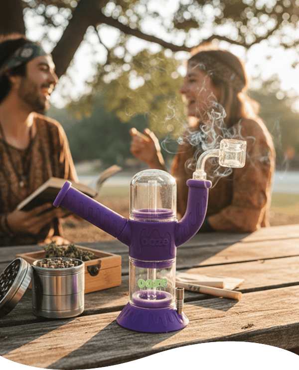 Close-up of the scarlet Ooze Cranium Silicone 4-in-1 Hybrid Bong on a picnic table with smoke curling from its glass joint and two people blurred in the sunlit background