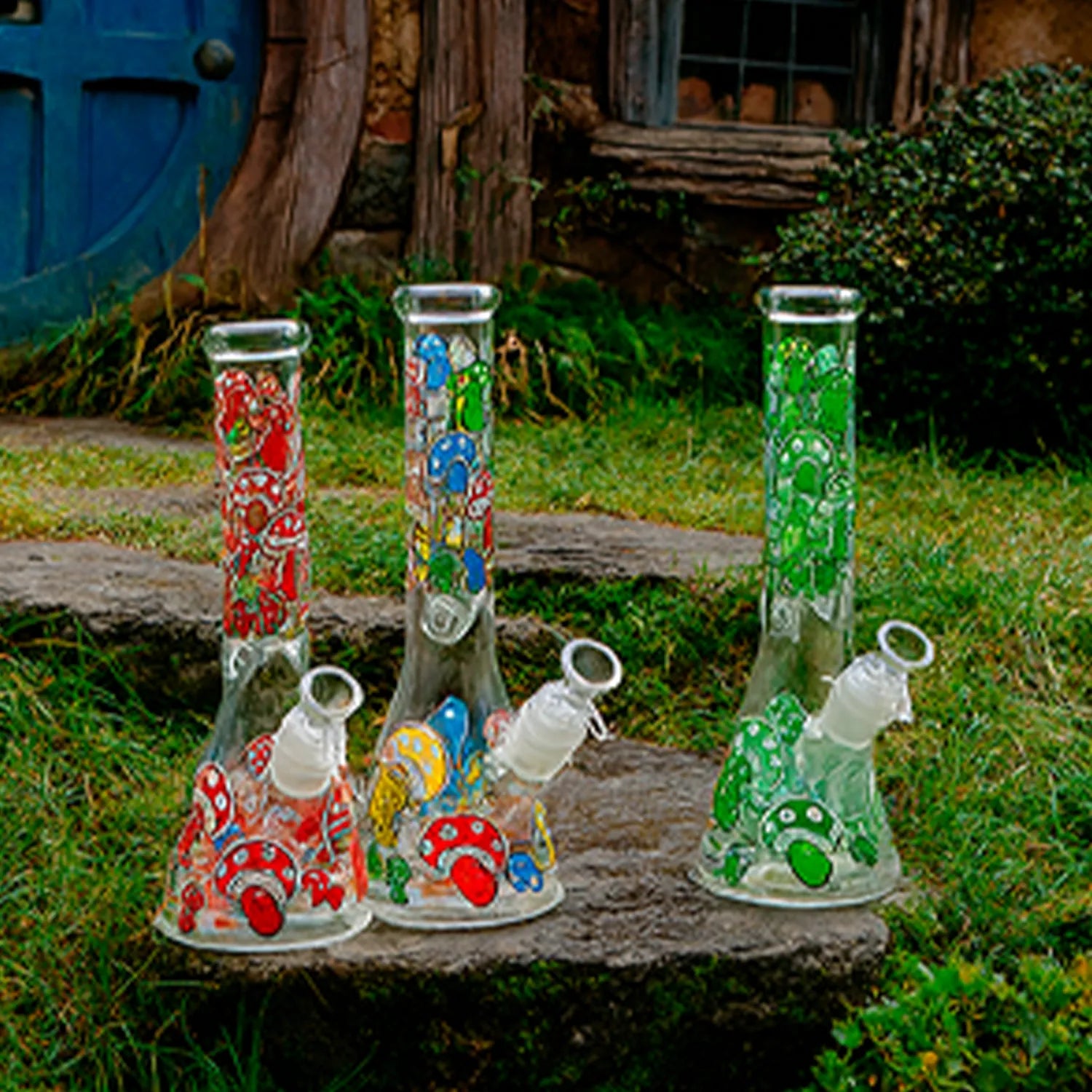 Three-quarter front view of the Glow Mushroom 10" Beaker Bong showing three colorful mushroom-decorated glass bongs on stone steps in a grassy garden.