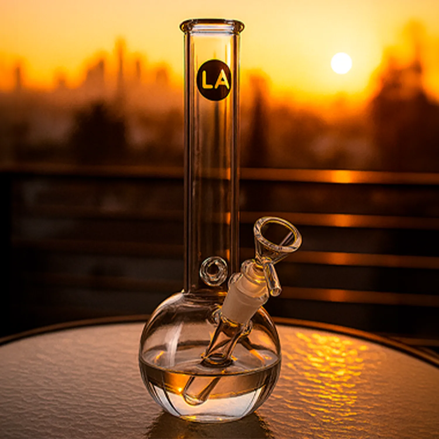 Sunlit close-up of the "Baller" Bubble Base Bong standing upright on a table with a round bubble base and city skyline blurred at sunset.