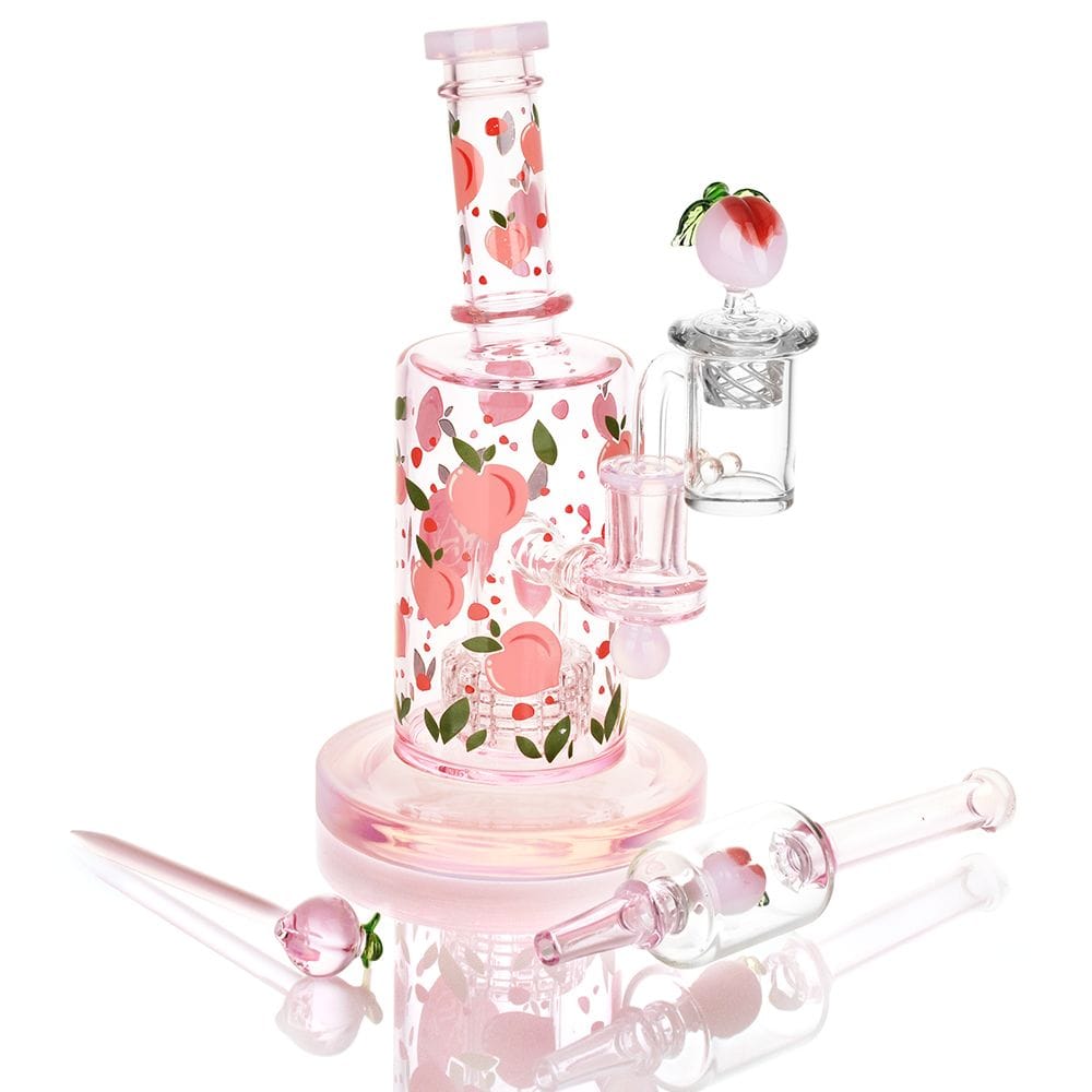 Angled front view of the Pulsar Fruit Series Peaches & Cream Glow In The Dark Dab Rig Kit - 8" / 14Mm F featuring pink glass with peach decals, matching attachments, and reflective white background