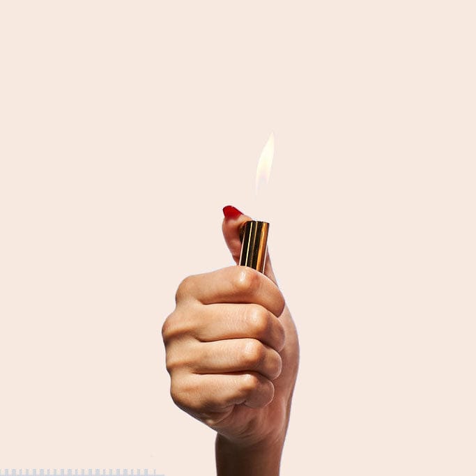 A close-up, low-angle view shows a hand holding the lit The De-Lighter, with a warm, golden flame.
