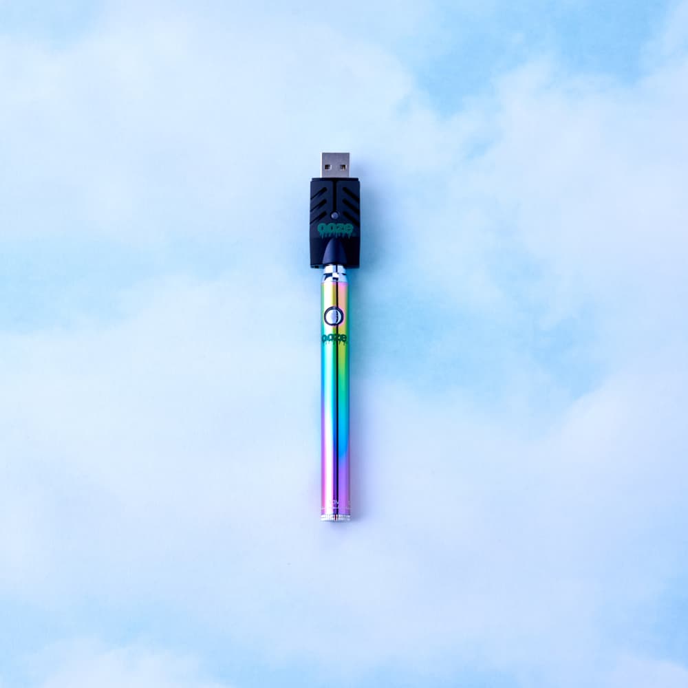 Top-down view of the black Ooze Slim Twist 510 Thread 320 mAh CBD Vape Pen Battery + USB Charger with a rainbow battery shaft against a soft blue cloudy background