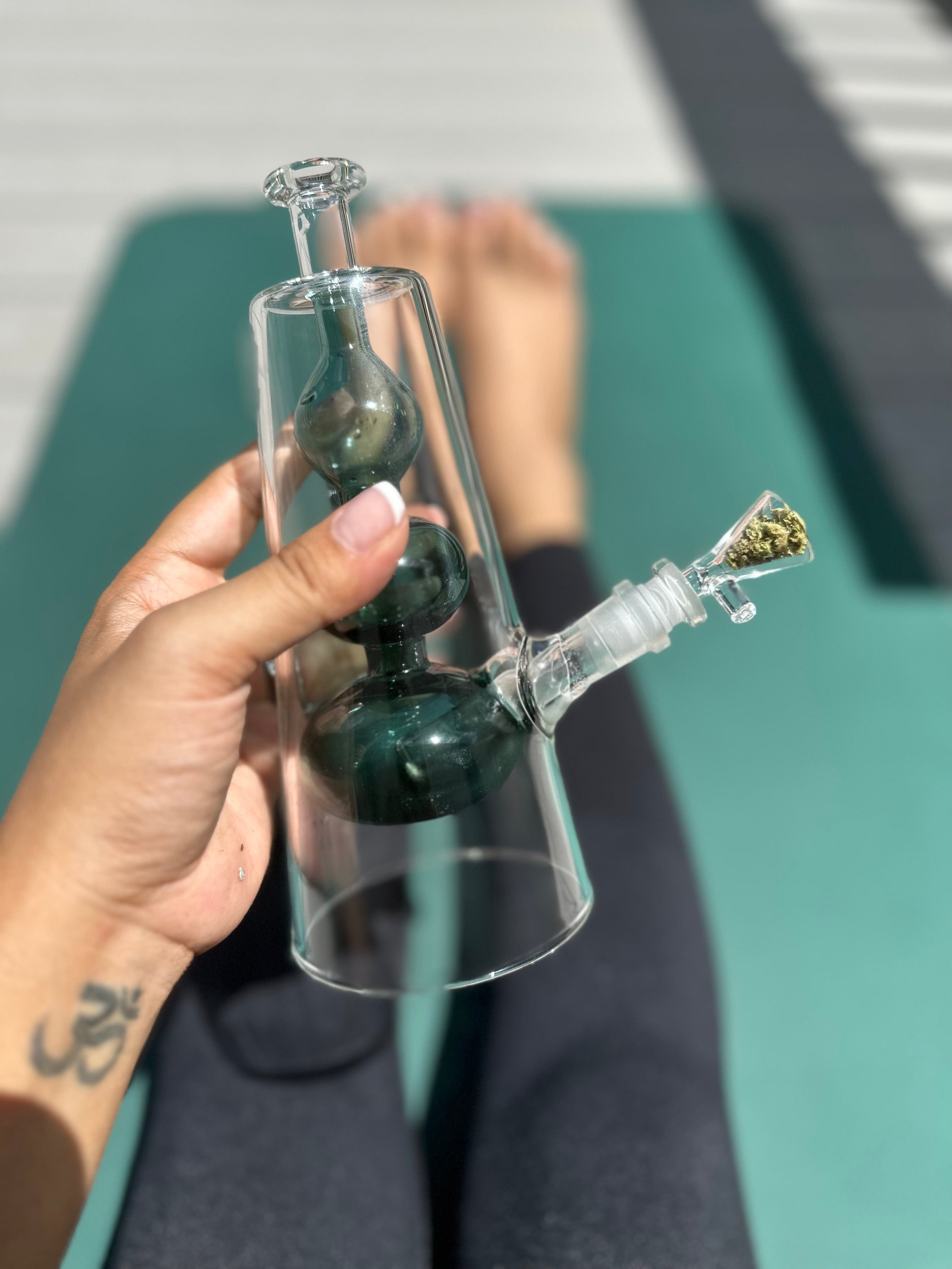 A close-up, angled view of The Bubblah, showing its clear glass construction held against a teal background.