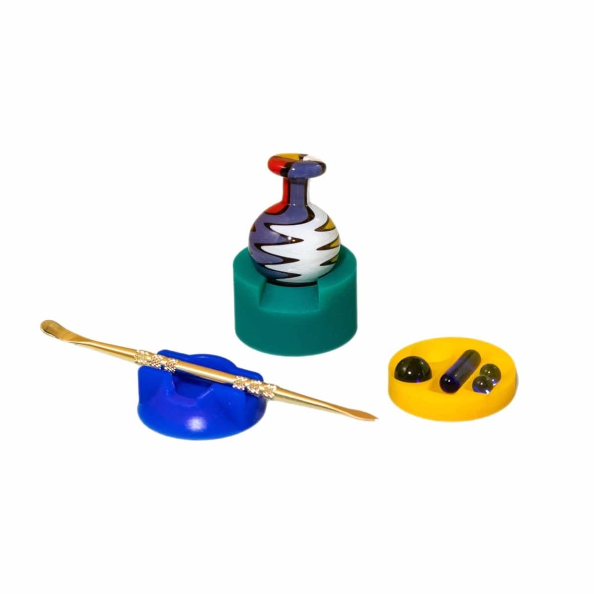 Center-focused product shot of the New Drop 3-pack *includes dabber* showing a striped glass carb cap perched on a green foam stand, a gold dabber resting on a blue holder, and a yellow tray with glass marbles.